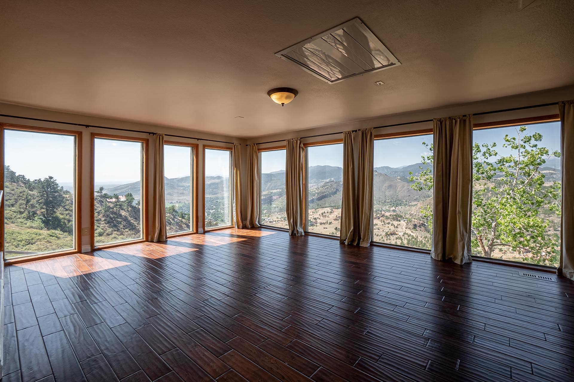 Room with floor-to-ceiling windows offering a mountain landscape view, dark wood flooring, and beige curtains framing the scenery.