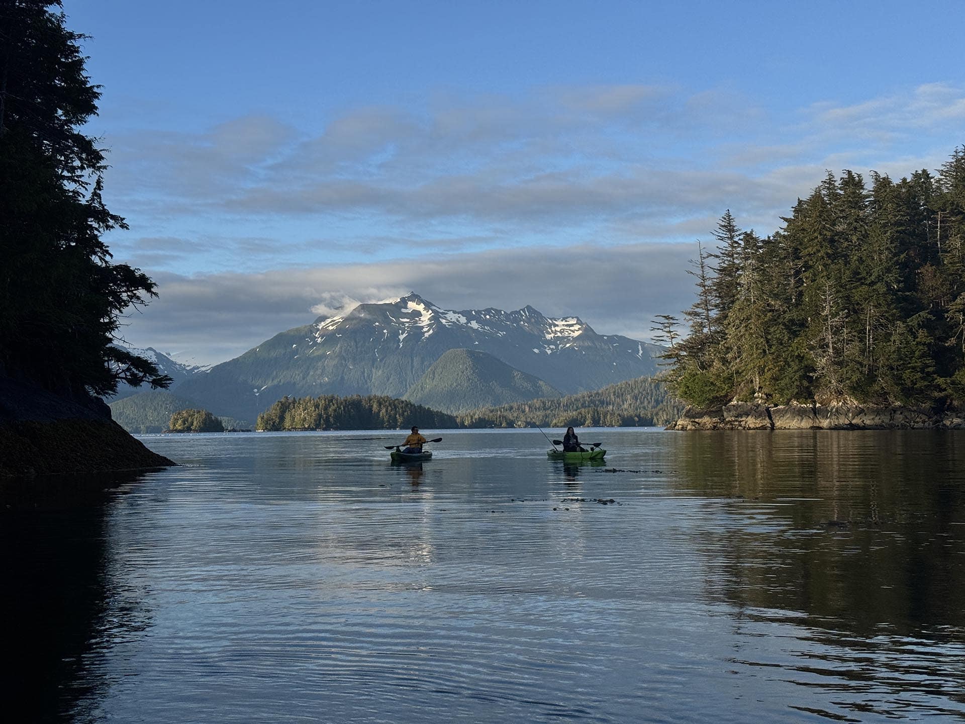 Two kayakers paddle on a calm lake with forested shores and snow-capped mountains under a blue sky.