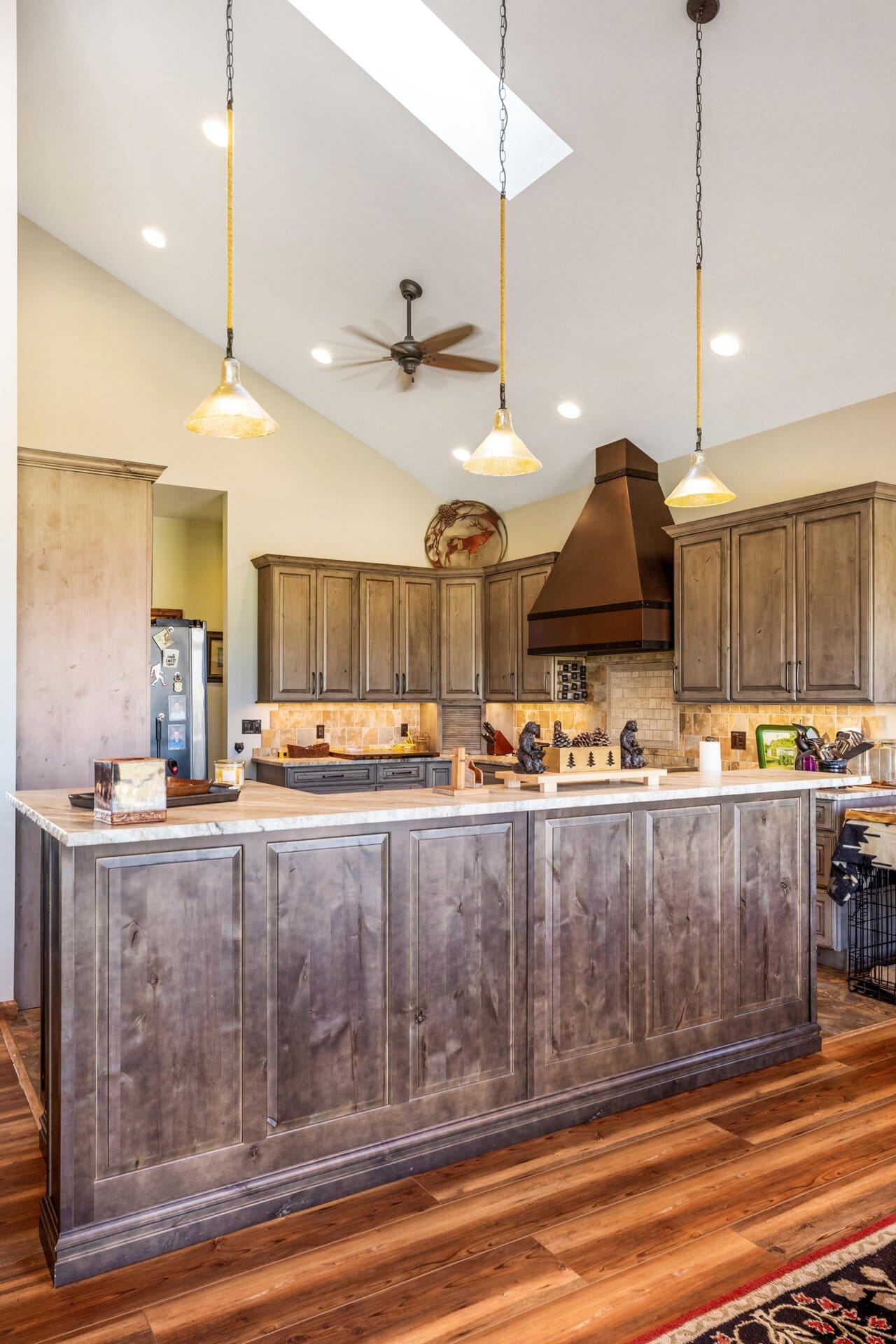 Open-concept kitchen with a long wooden island, pendant lights, and a copper vent hood above the stove. Cabinets are a warm gray-brown; wood floor visible.