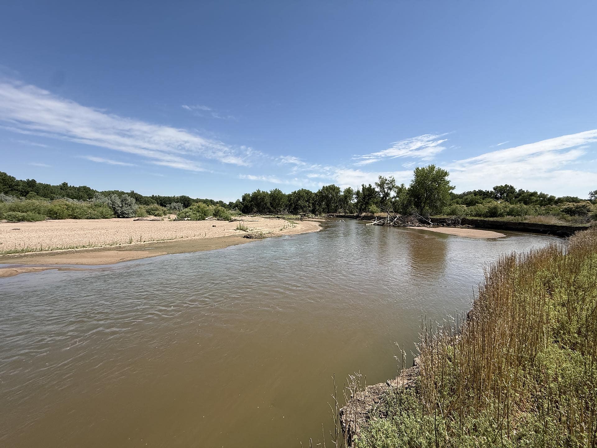 Muddy river with sandy banks and green trees along the shore under a blue sky.