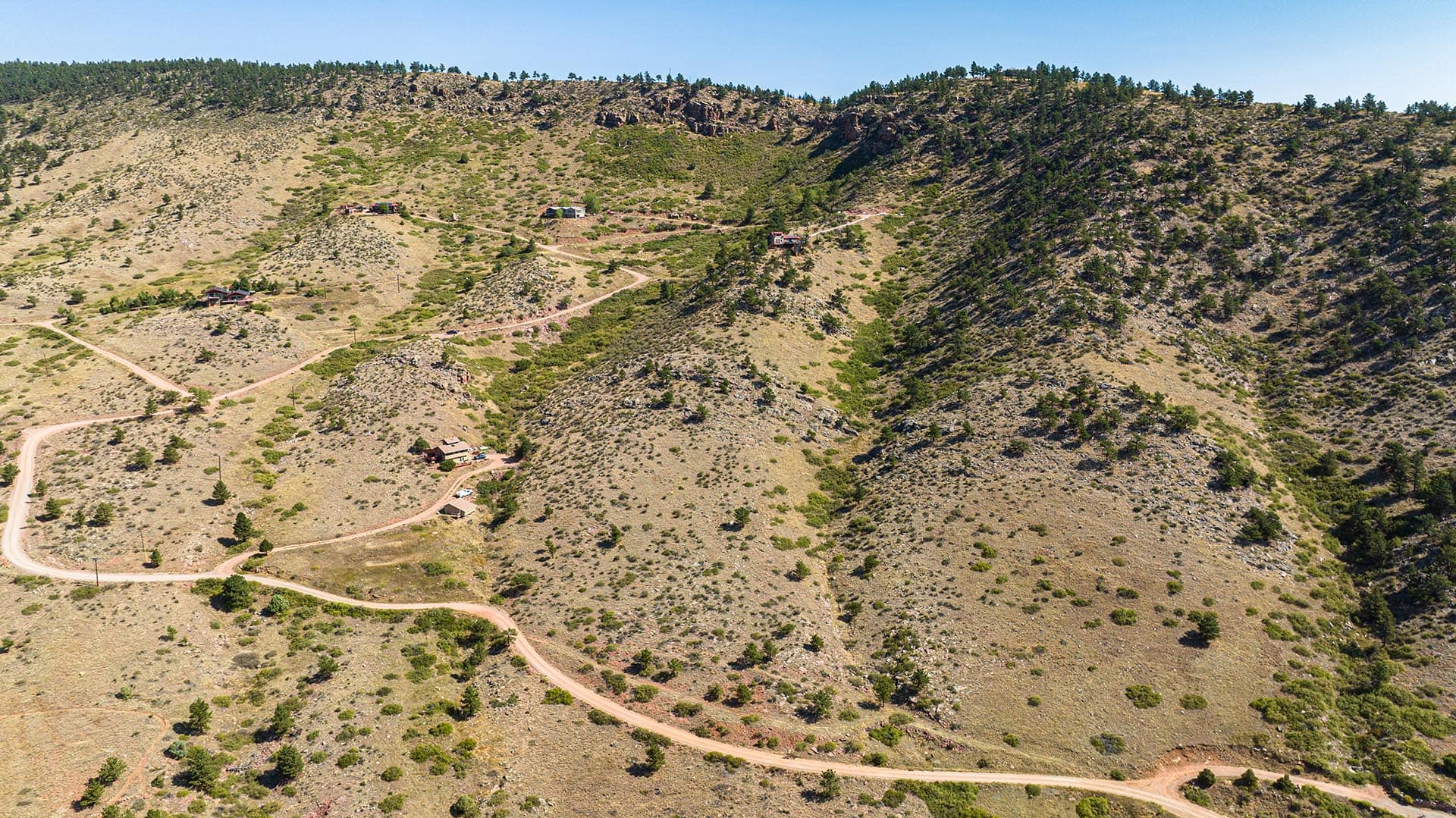 Aerial view of a dry, hilly landscape with dirt roads winding through scattered houses and sparse trees under a clear blue sky.