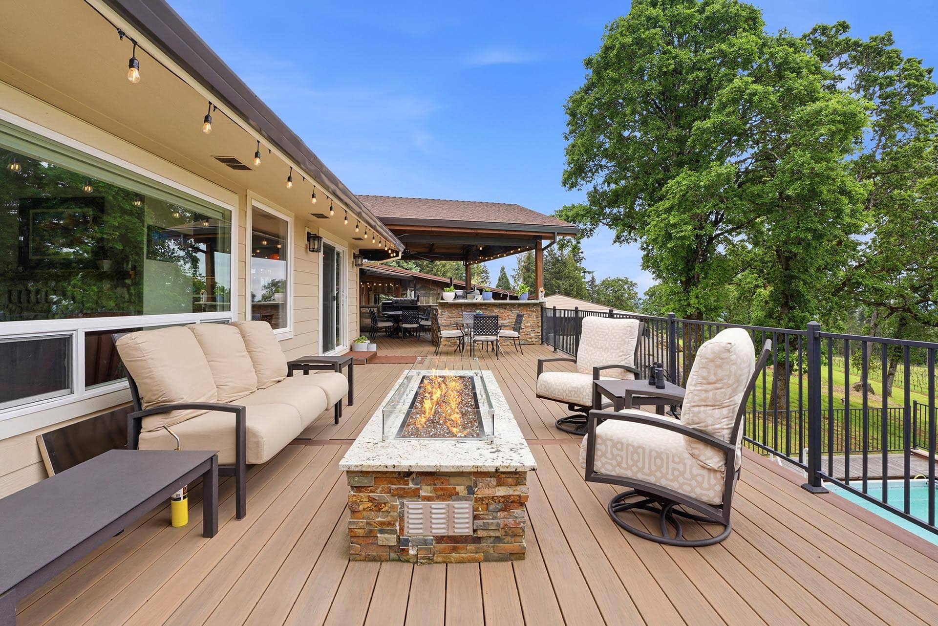 Outdoor deck with beige cushioned seating around a stone-firepit table, string lights, and a railing; large tree and blue sky in background.