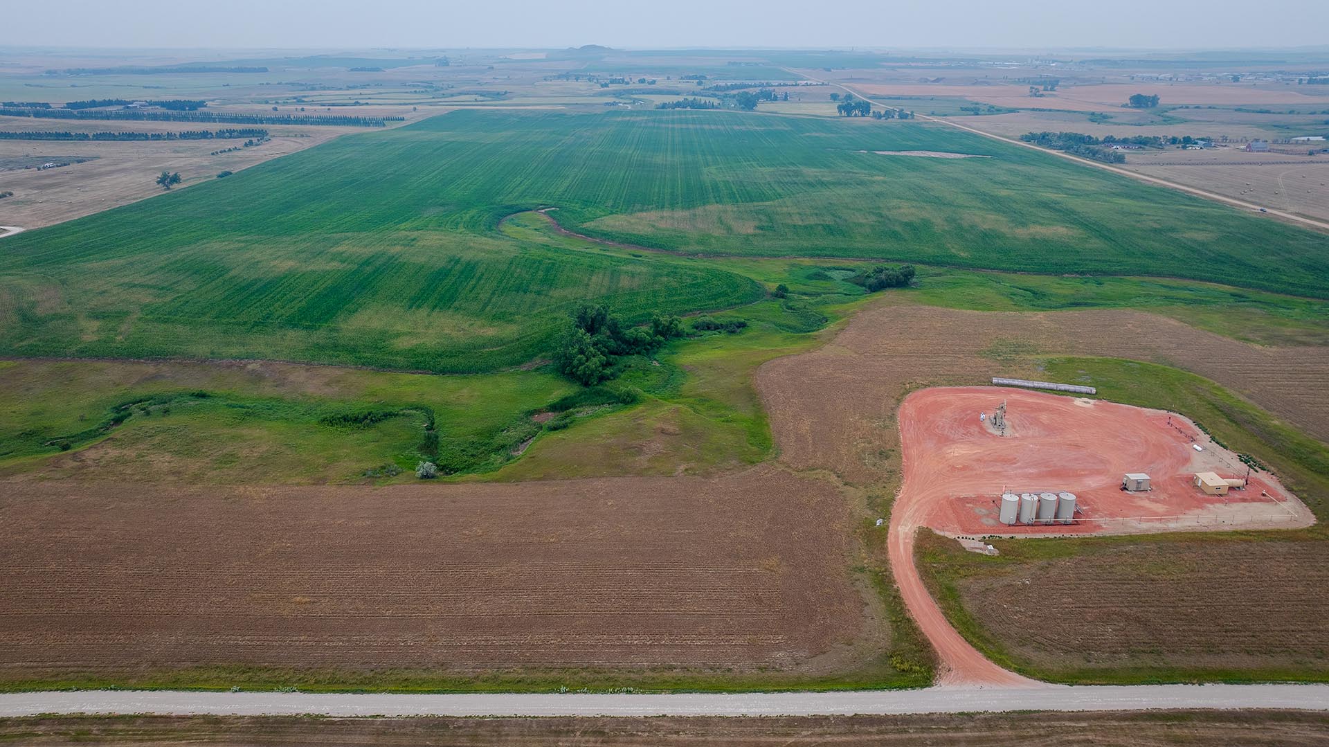 Aerial view of green fields and brown farmland with a red-dirt development pad and silos at a construction site in the foreground.