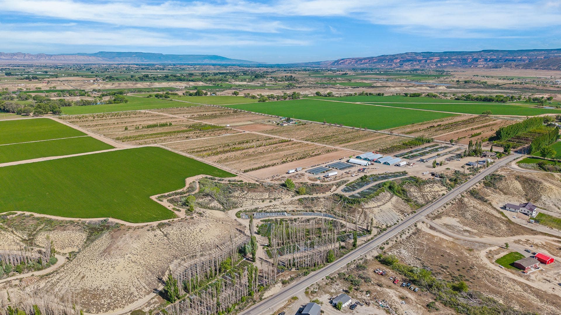 Aerial view of farmland with green fields, dirt roads, and a farm complex near a paved highway; distant mountains in the background.