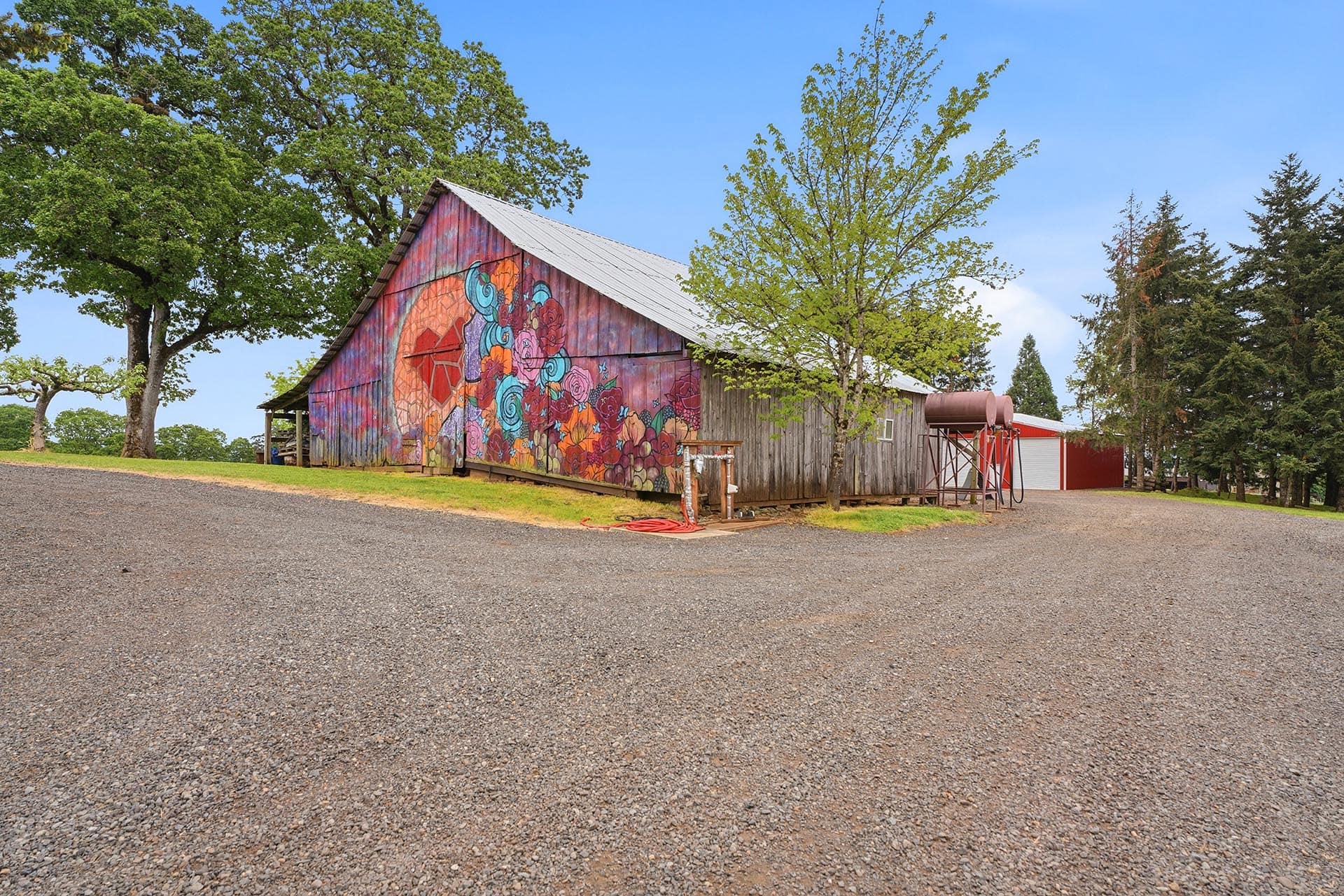 Old wooden barn with a bright floral mural and a metal roof, set in a gravel lot with green trees and a clear blue sky behind it.