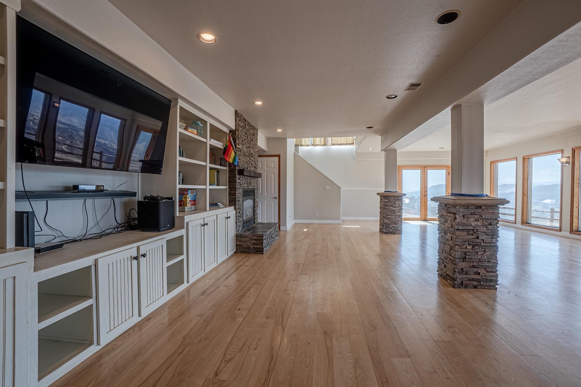 Spacious living area with built‑in white cabinets, a wall-mounted TV, and a stone fireplace, overlooking mountains through large windows.