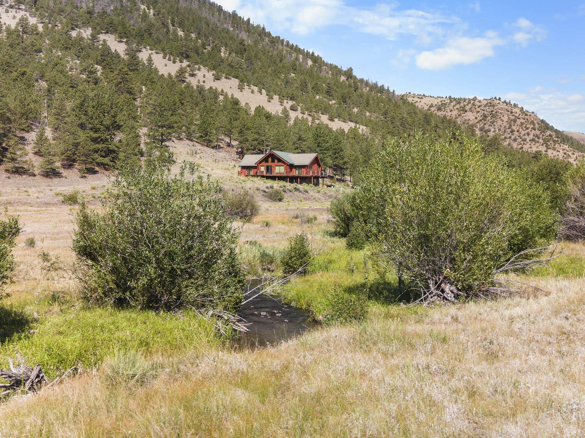 Red wooden cabin on stilts beside a narrow stream, surrounded by dry grass and pine-covered hills under a bright blue sky.
