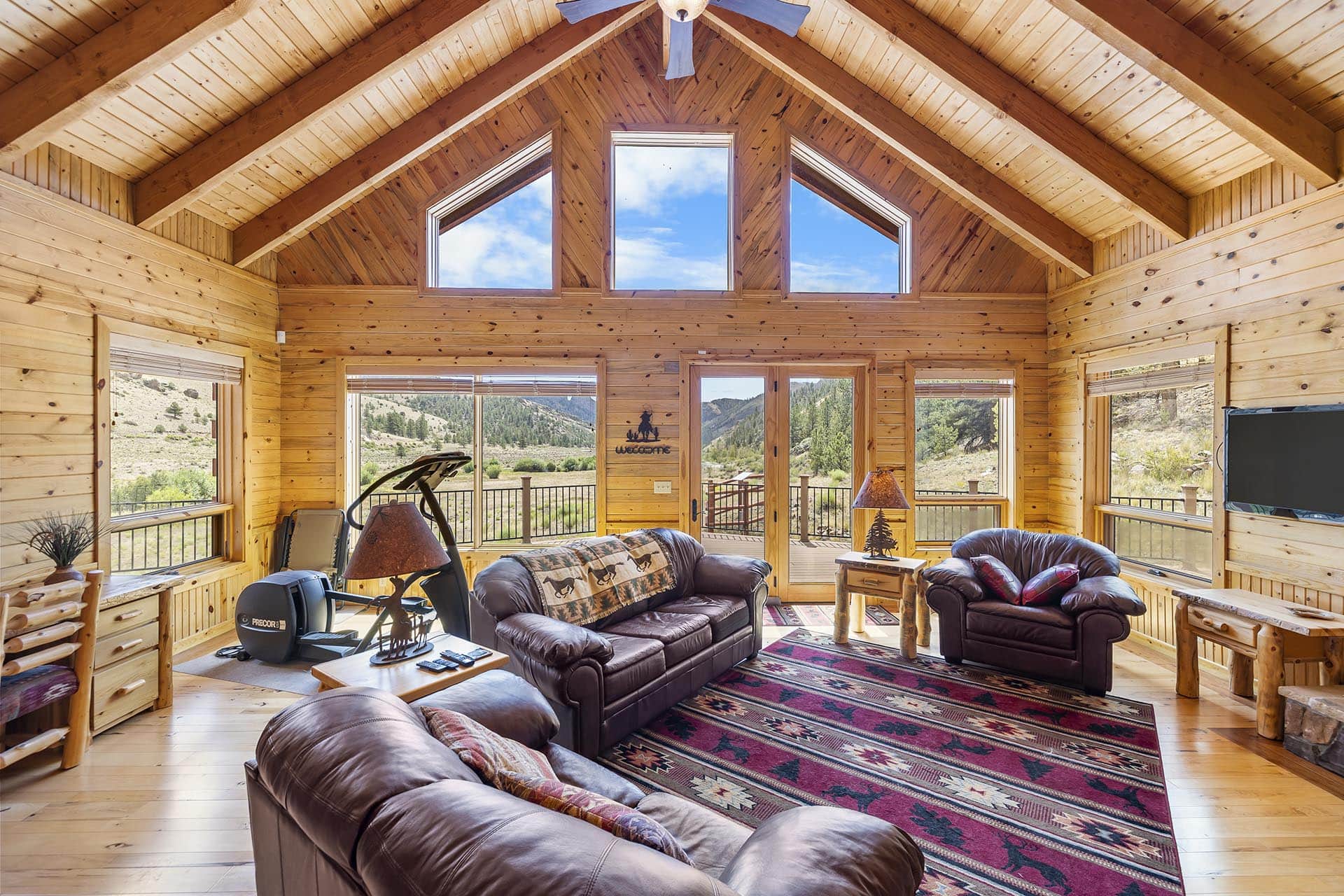 Cozy log-cabin living room with a vaulted wood ceiling and large triangular windows showing blue sky and mountains outside.