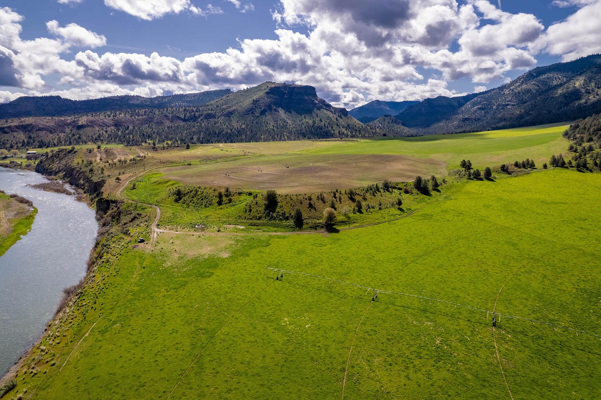 Aerial view of a river winding through green fields toward distant mountains under a cloudy blue sky.
