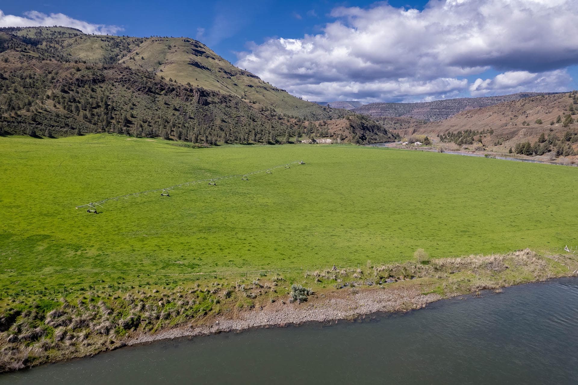 A wide valley with green pasture, a winding river, and hills in the distance under a blue sky with clouds; center-pivot irrigation runs across the field.