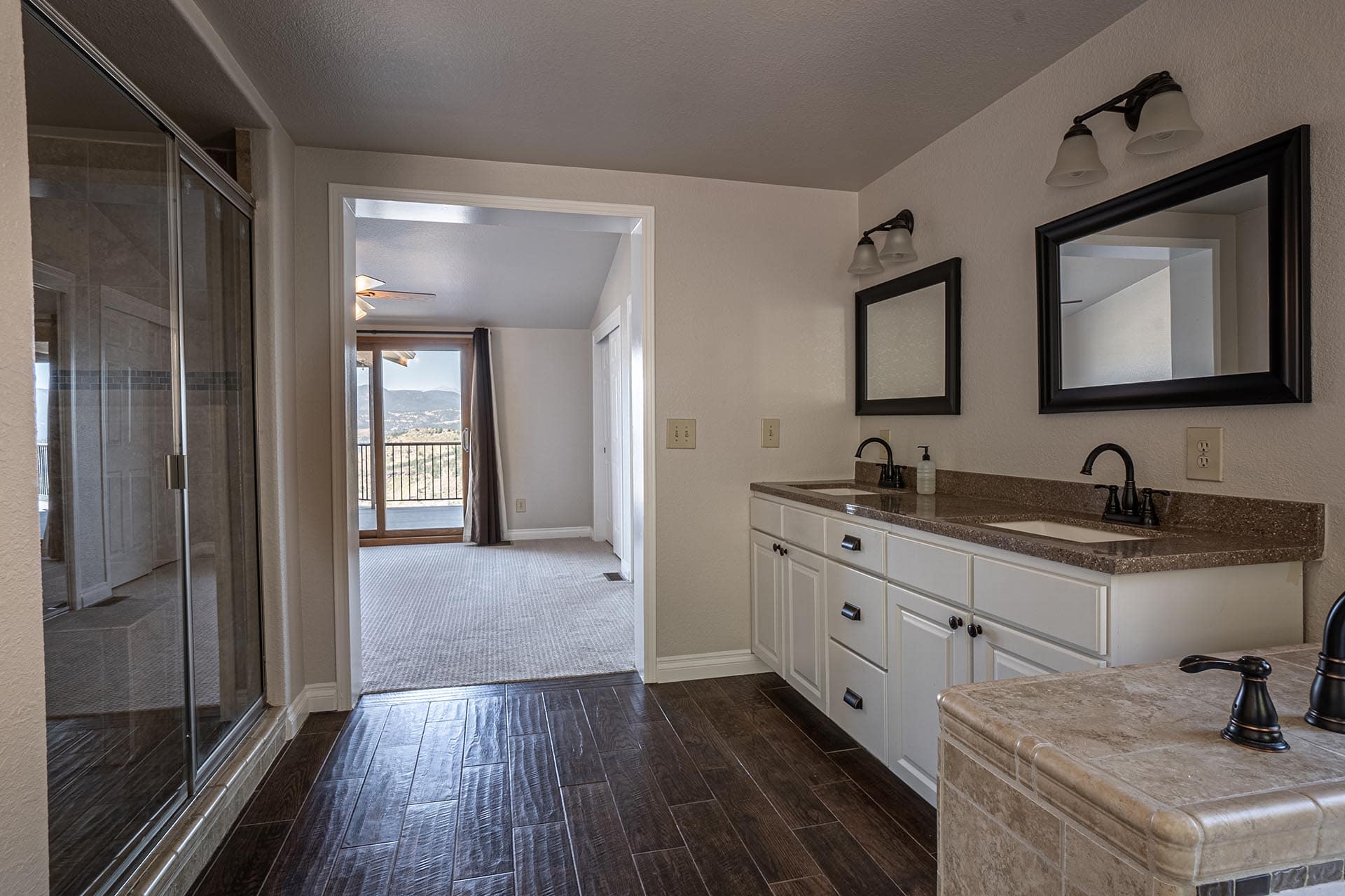 Bathroom with double sink vanity, two mirrors, and a glass shower; doorway opens to a room with a balcony and view of mountains.
