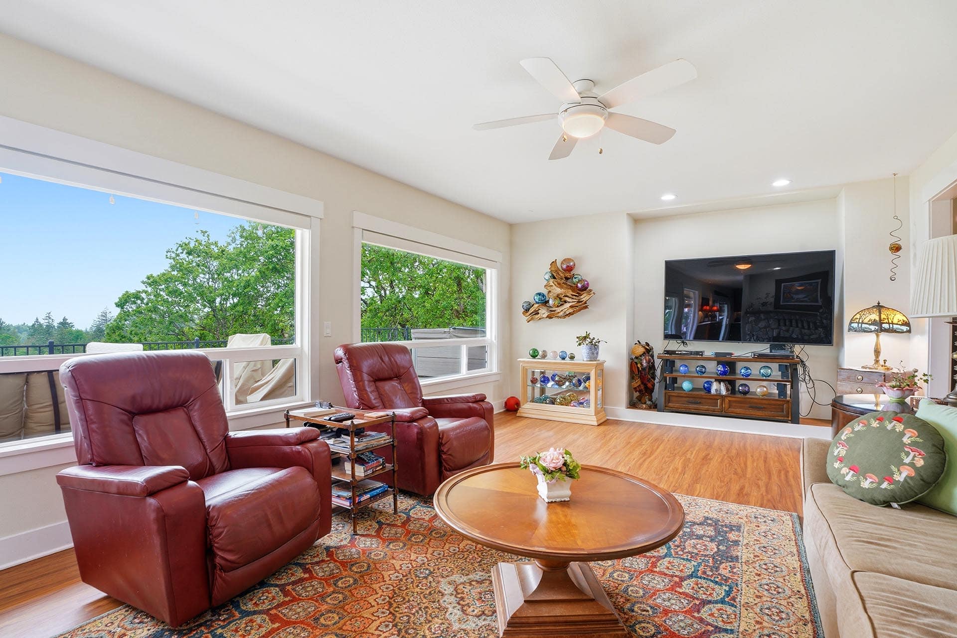 Bright living room with red leather recliners, a round coffee table, and a wall-mounted TV; large windows show trees outside. Avoids glare.