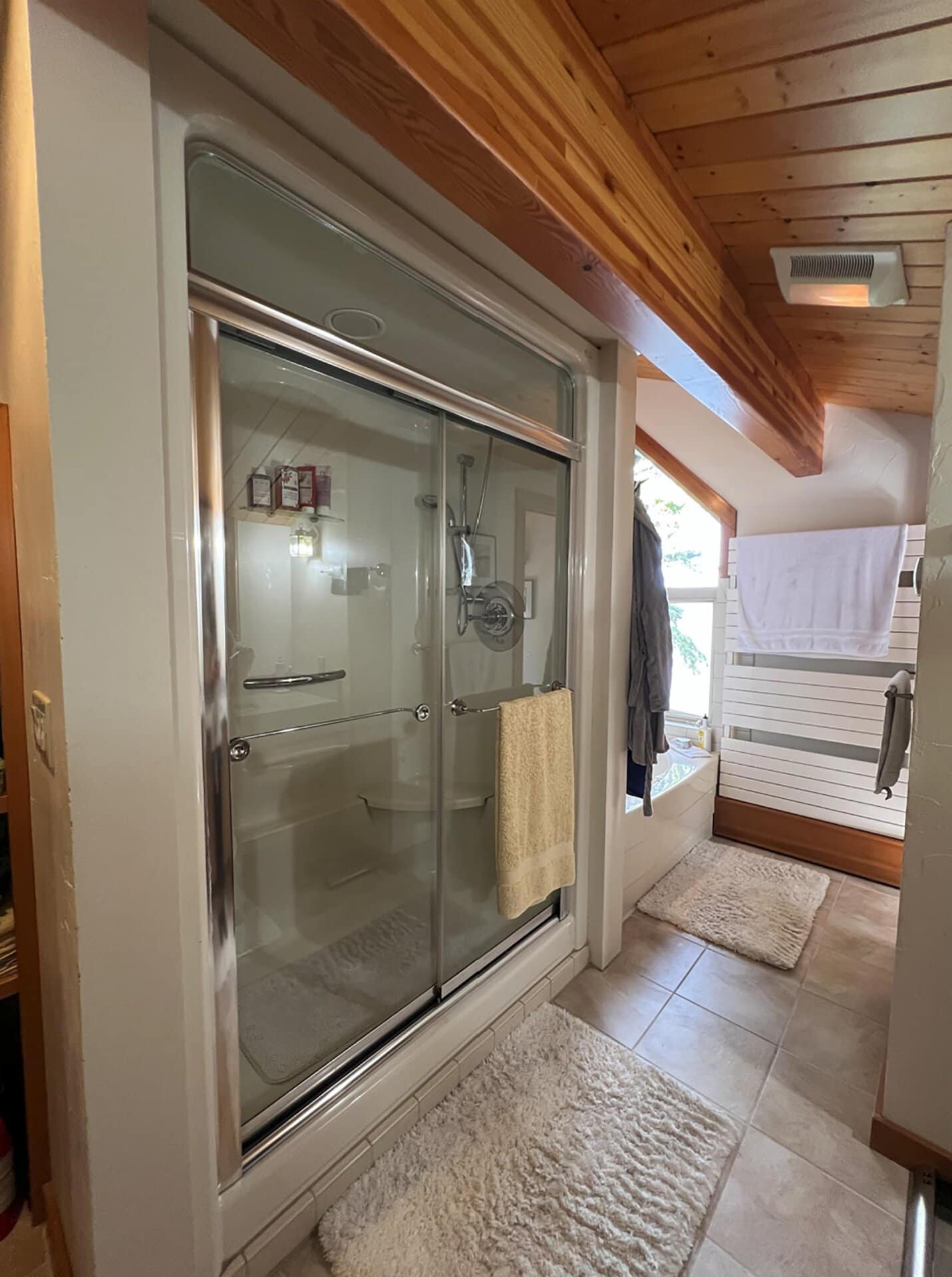 Glass-enclosed shower in a rustic bathroom with a wood-beamed ceiling, white towels, and fluffy white bath mats on a tiled floor.