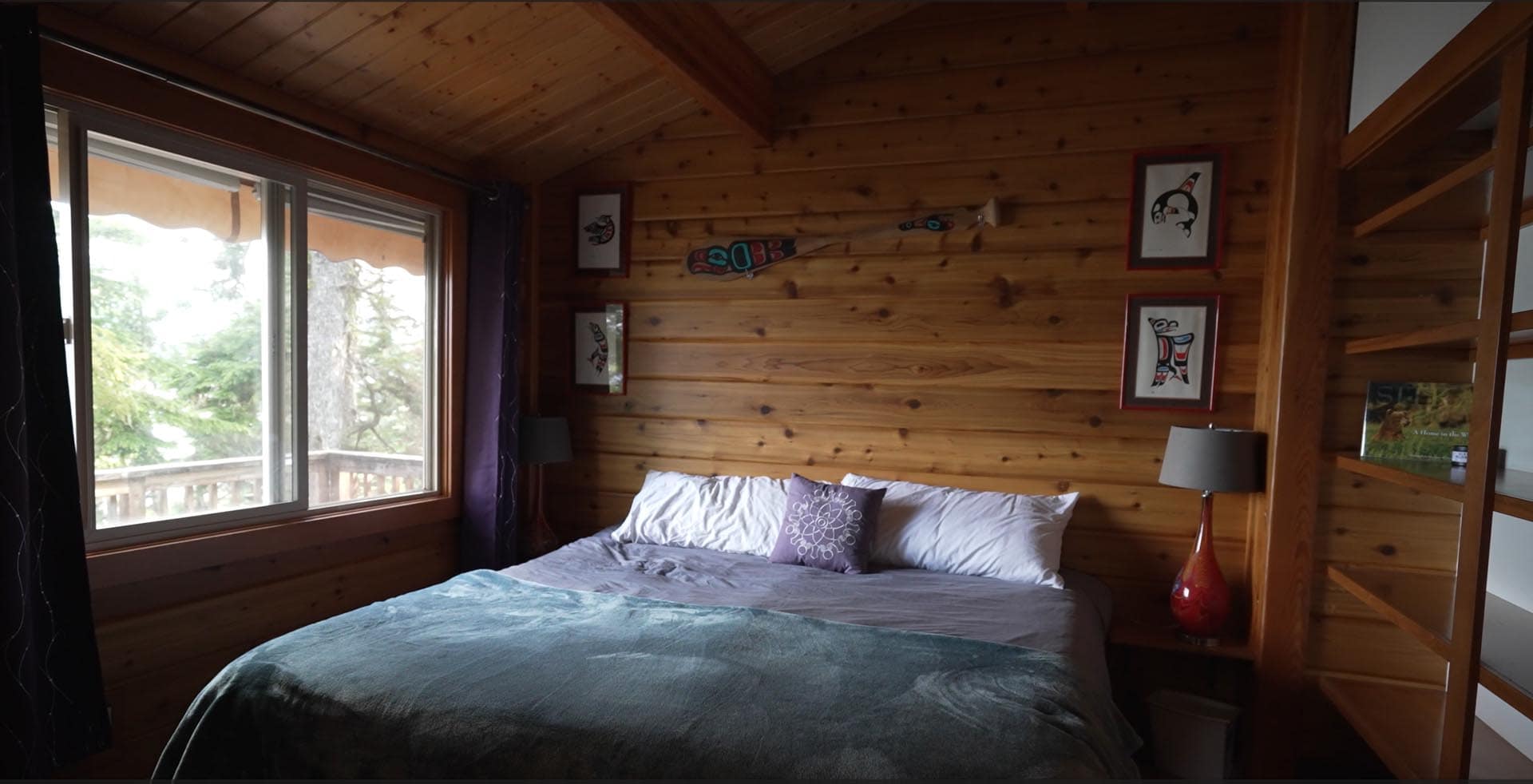 Cozy cabin bedroom with knotty pine walls, a large bed, white and purple pillows, and a window to a deck outside.