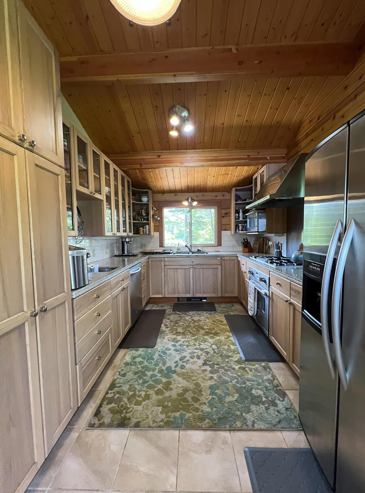 Rustic galley kitchen with light wood cabinets, stainless appliances, and a green floral rug running down the center.