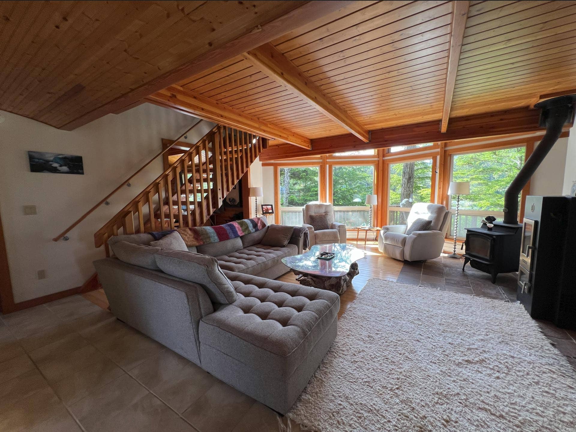 Cozy living room with a gray sectional, glass coffee table, and large windows showing trees outside.