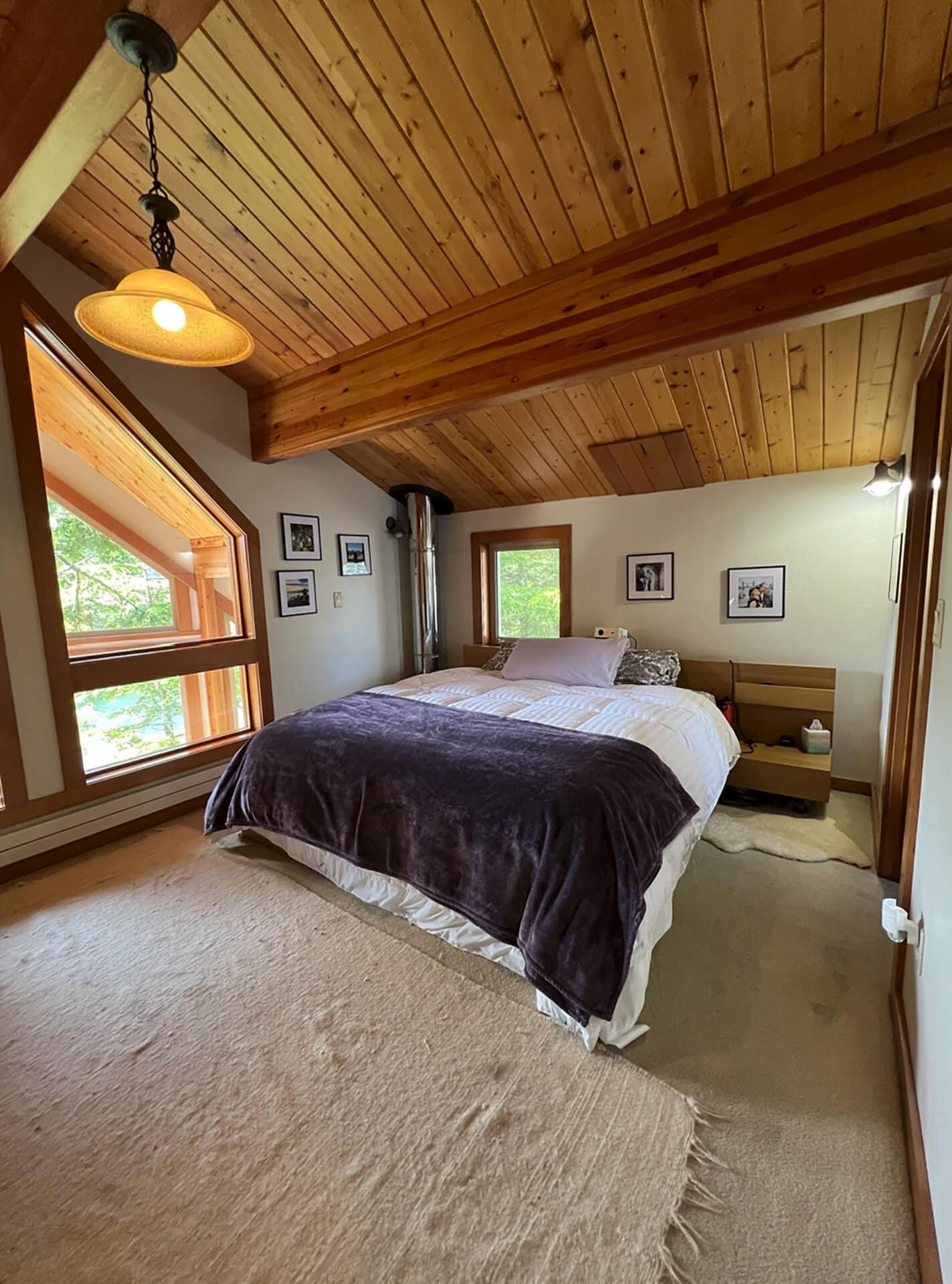 Rustic bedroom with a sloped wooden ceiling, exposed beams, and a bed covered in white and dark throws, facing a window. Framed photos line the walls.