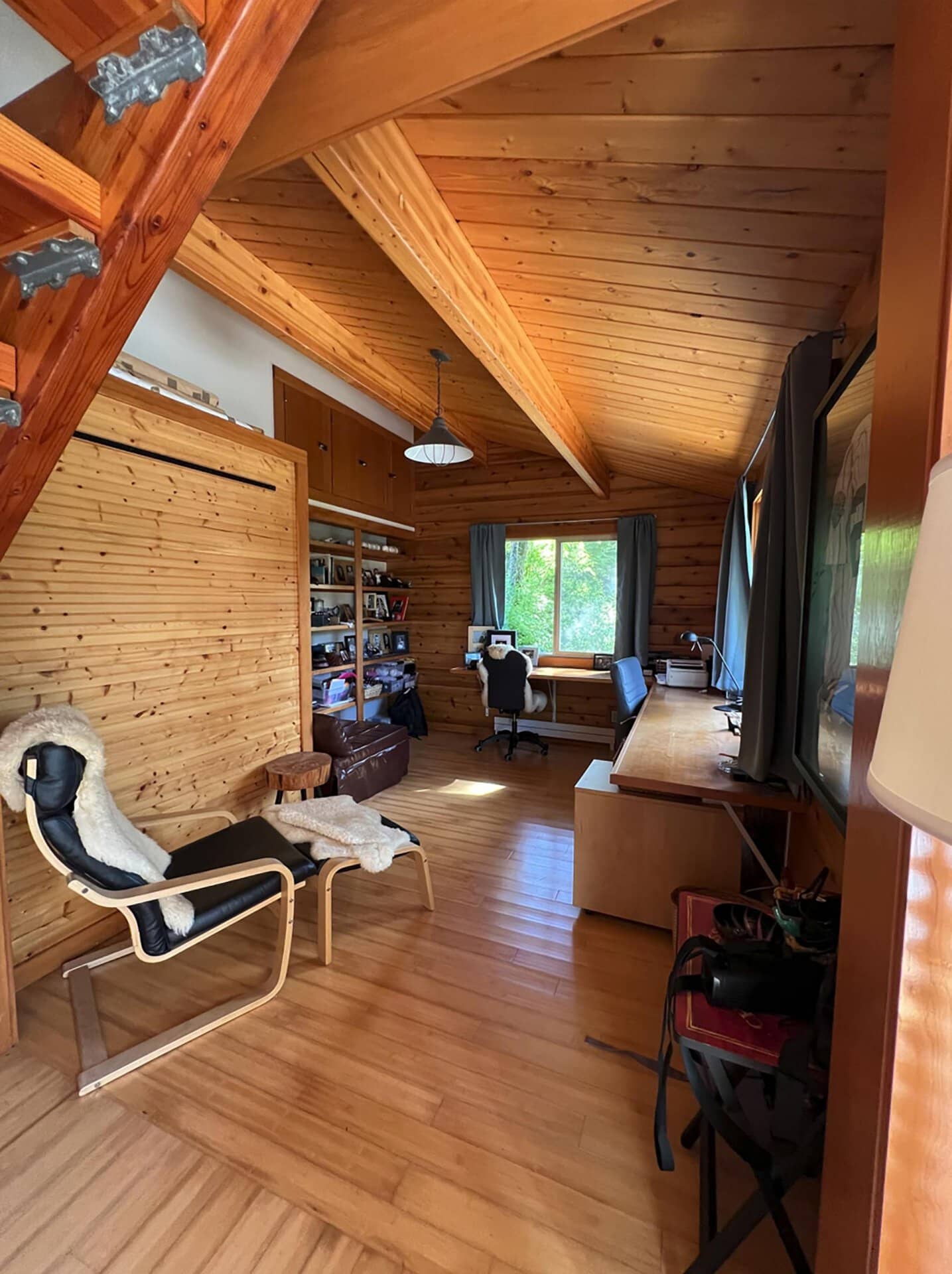 Cozy wood-paneled home office with slanted ceiling beams, desk by window, and bookshelves along the wall