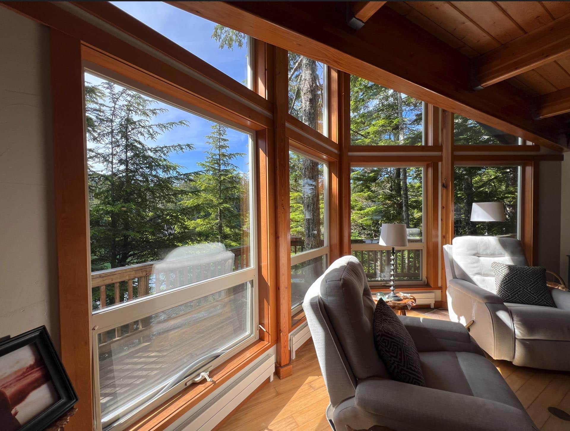 Sunlit living room with large wood-framed windows facing evergreen trees; gray armchairs and sofa indoors.