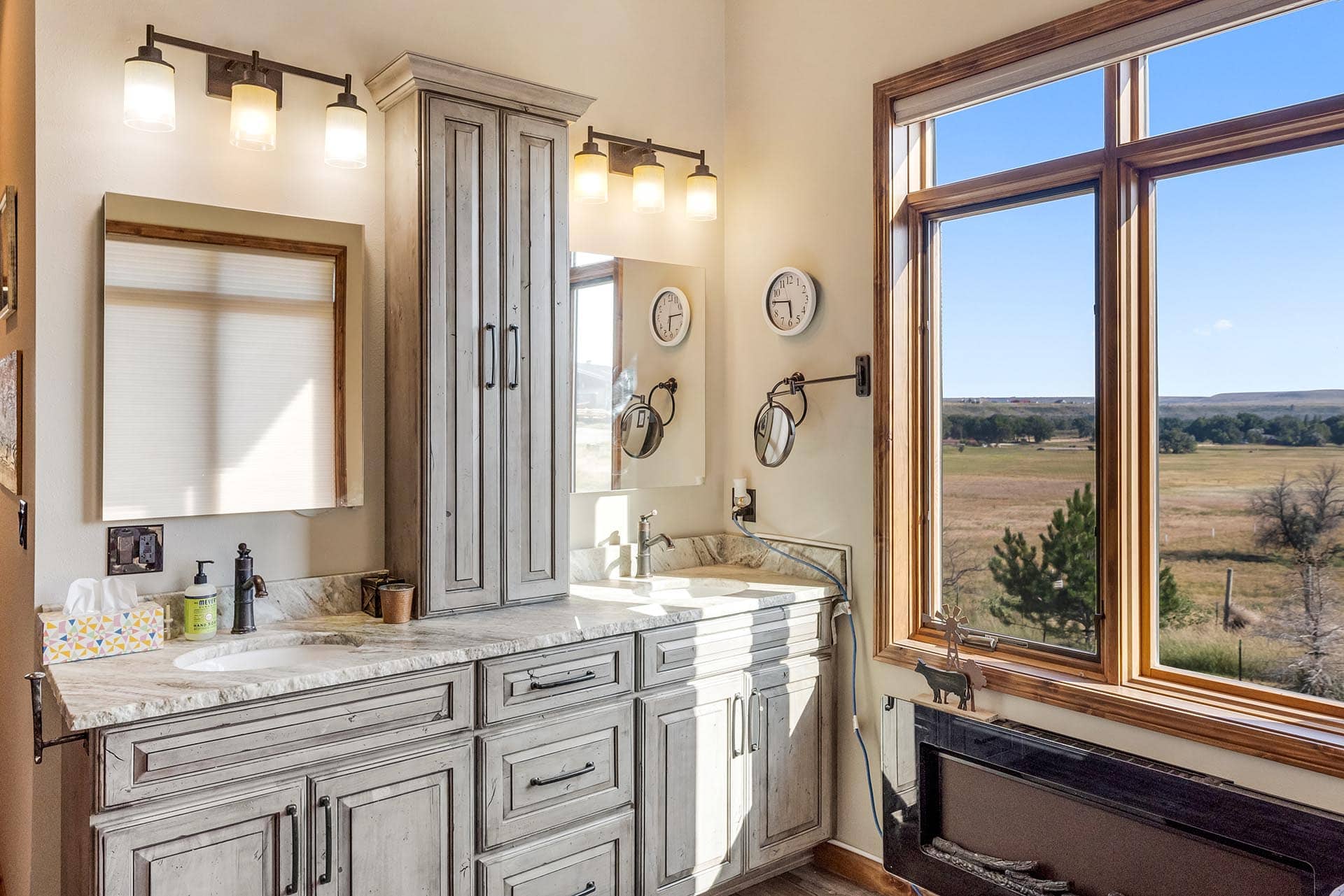 Grey distressed cabinetry and marble countertop bathroom vanity with two sinks and vertical mirrors by a large window overlooking a countryside landscape.