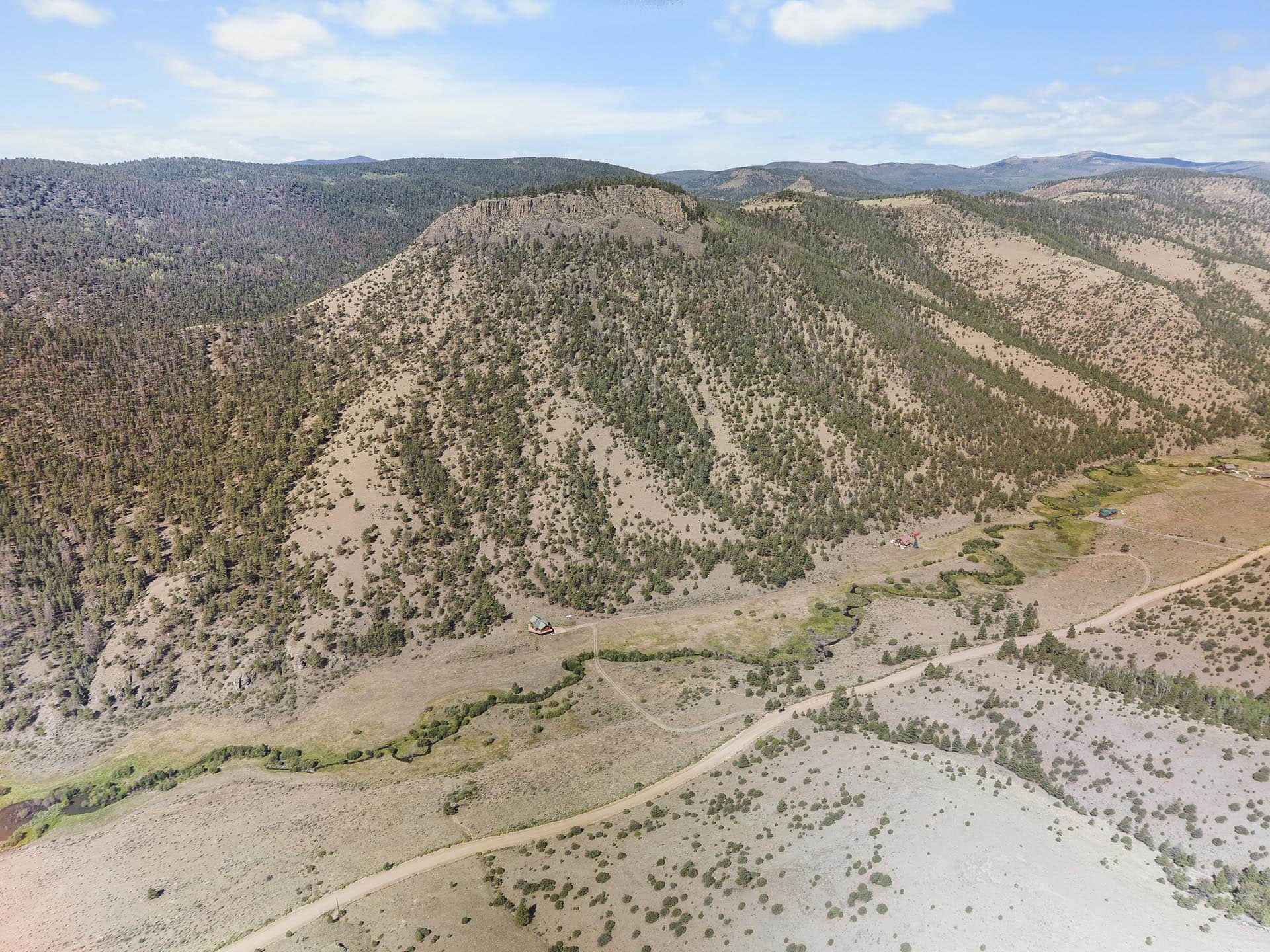 Aerial view of a dry, hilly landscape with scattered trees, winding dirt roads, and a few houses at the valley edge.