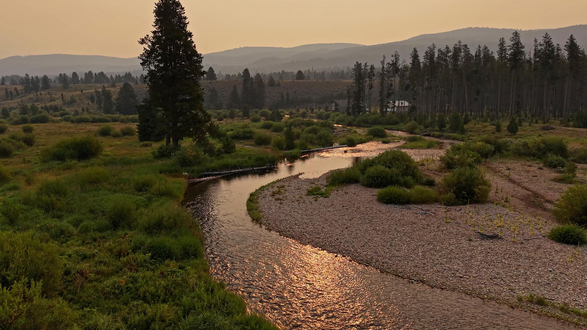 Sunset over a meandering river through a grassy valley with distant hills and a stand of trees on the right.