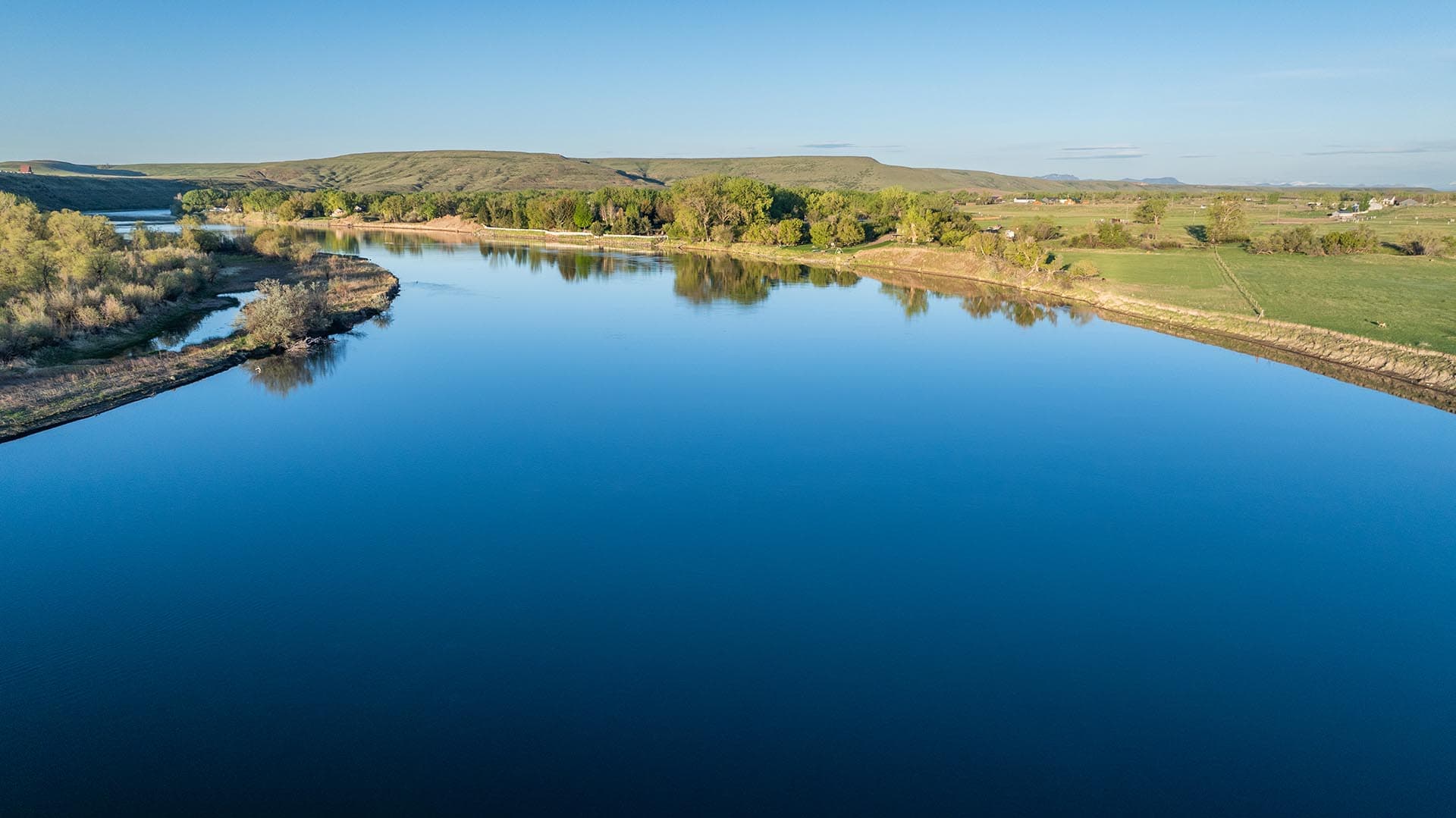 A calm river reflecting the blue sky, with tree-lined banks and green fields in a rural landscape.