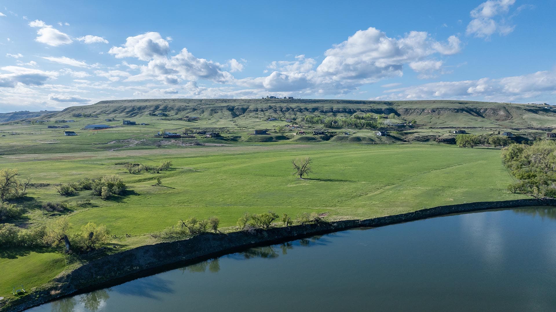 Green farmland with a gentle hill dotted by houses, and a calm river in the foreground beneath a blue, cloudy sky.