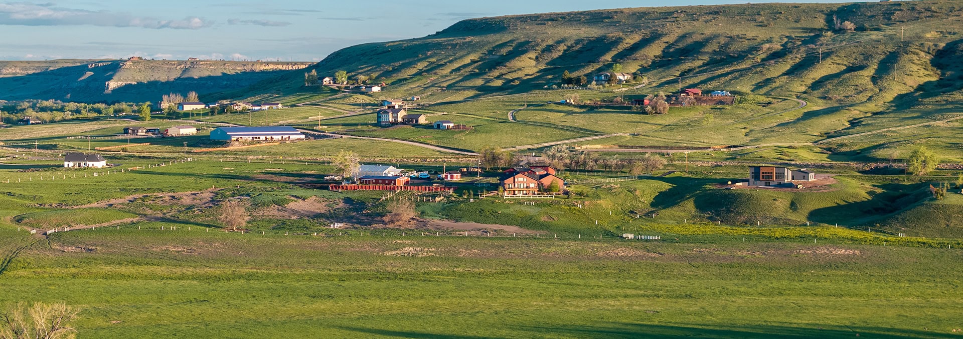 Wide rural hillside with scattered houses, green fields, and winding roads under a clear sky—a peaceful farm community seen from a distance.