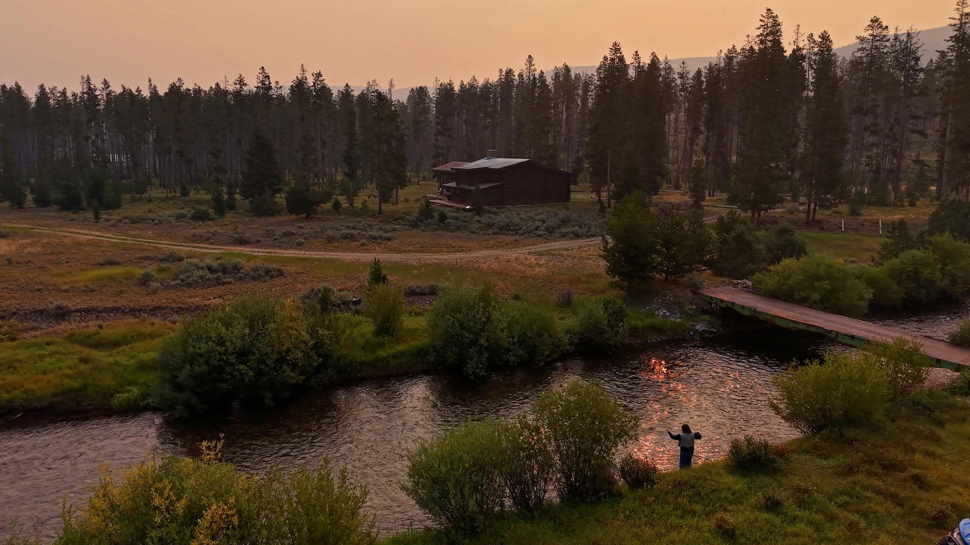 Log cabin in a pine forest by a winding river at sunset, with a wooden footbridge and a person standing by the water