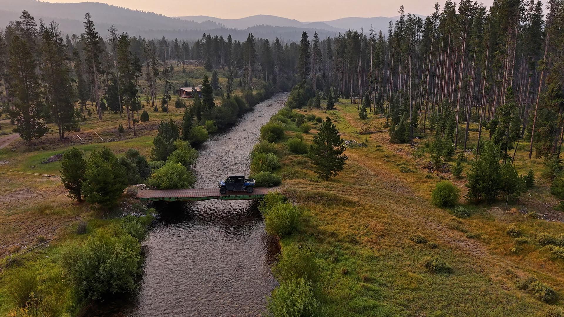 Aerial view of a river winding through a forested valley with a small wooden bridge and a jeep parked on it.