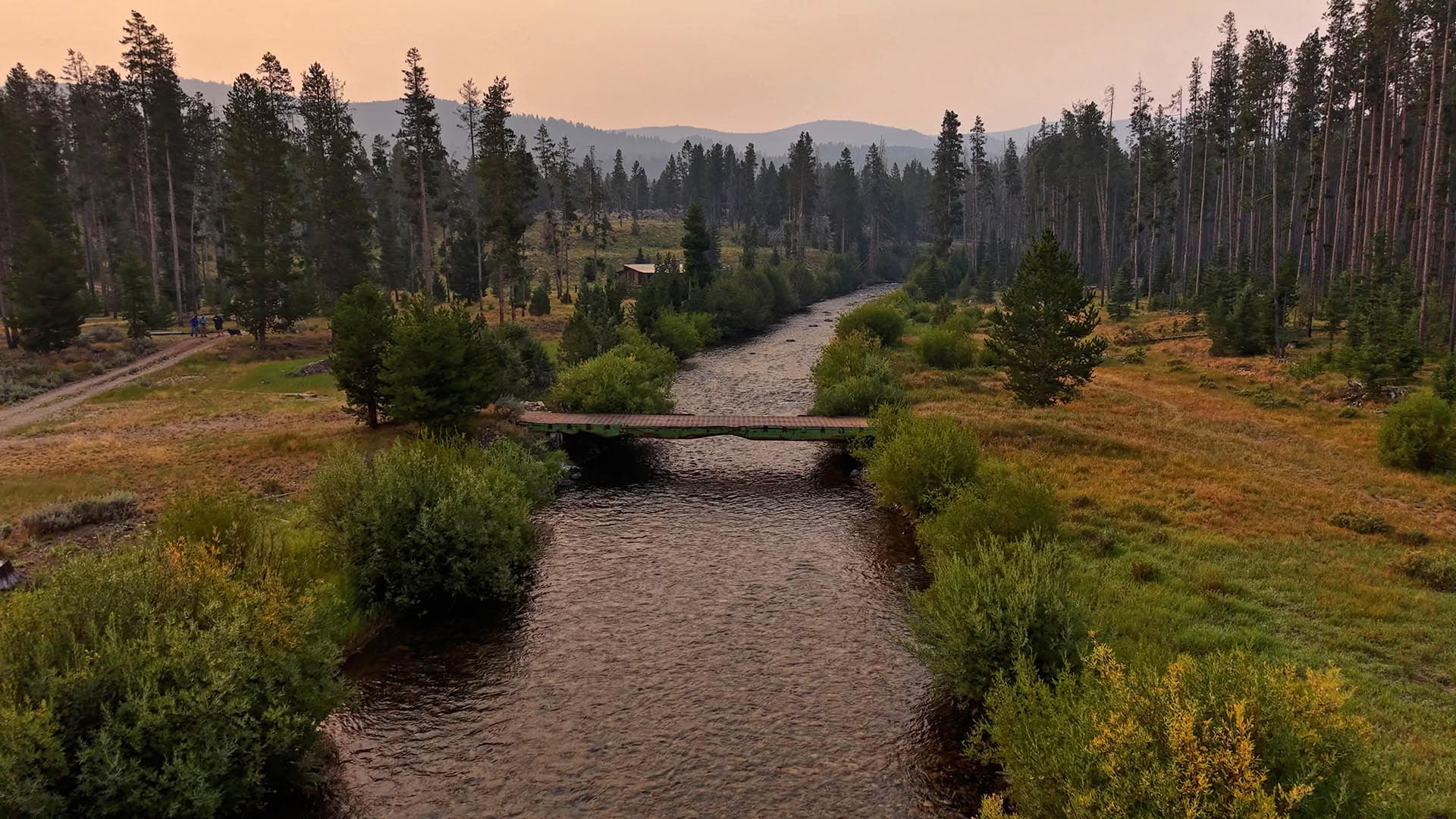 River winds through a forested valley with a small bridge crossing it at sunset.