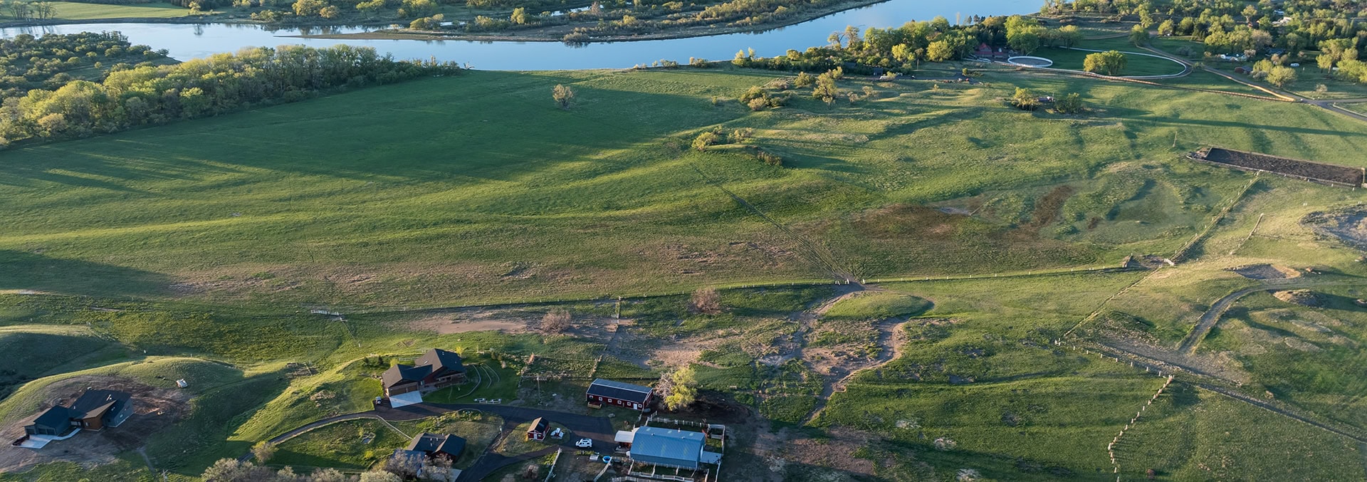 Aerial view of a rural landscape with green pastures, a winding river at the top, and farm buildings clustered in the bottom left.