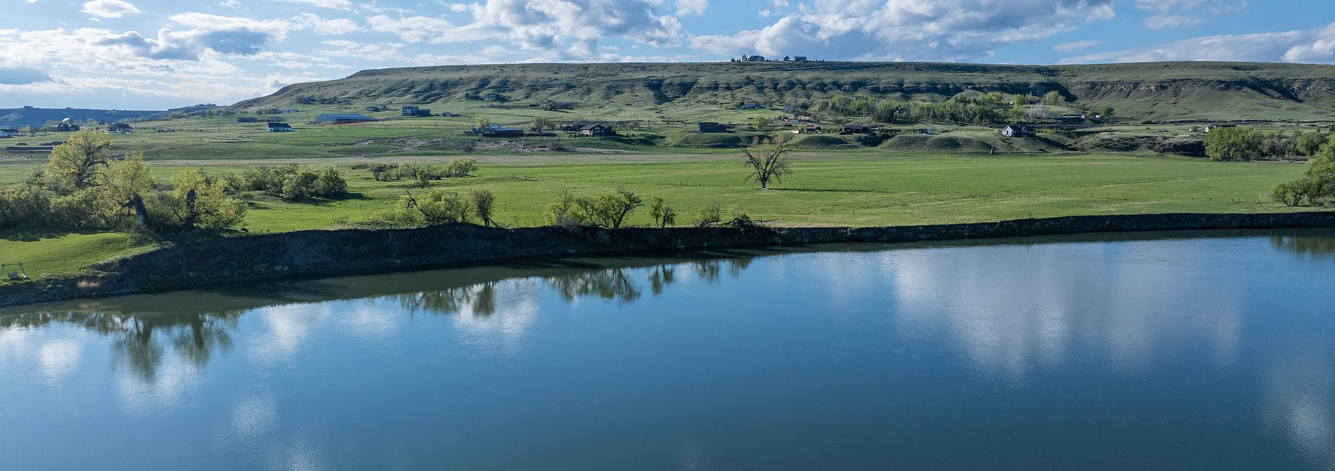 Wide river reflects a blue sky with clouds, green fields, and a low hill dotted with houses in the distance.