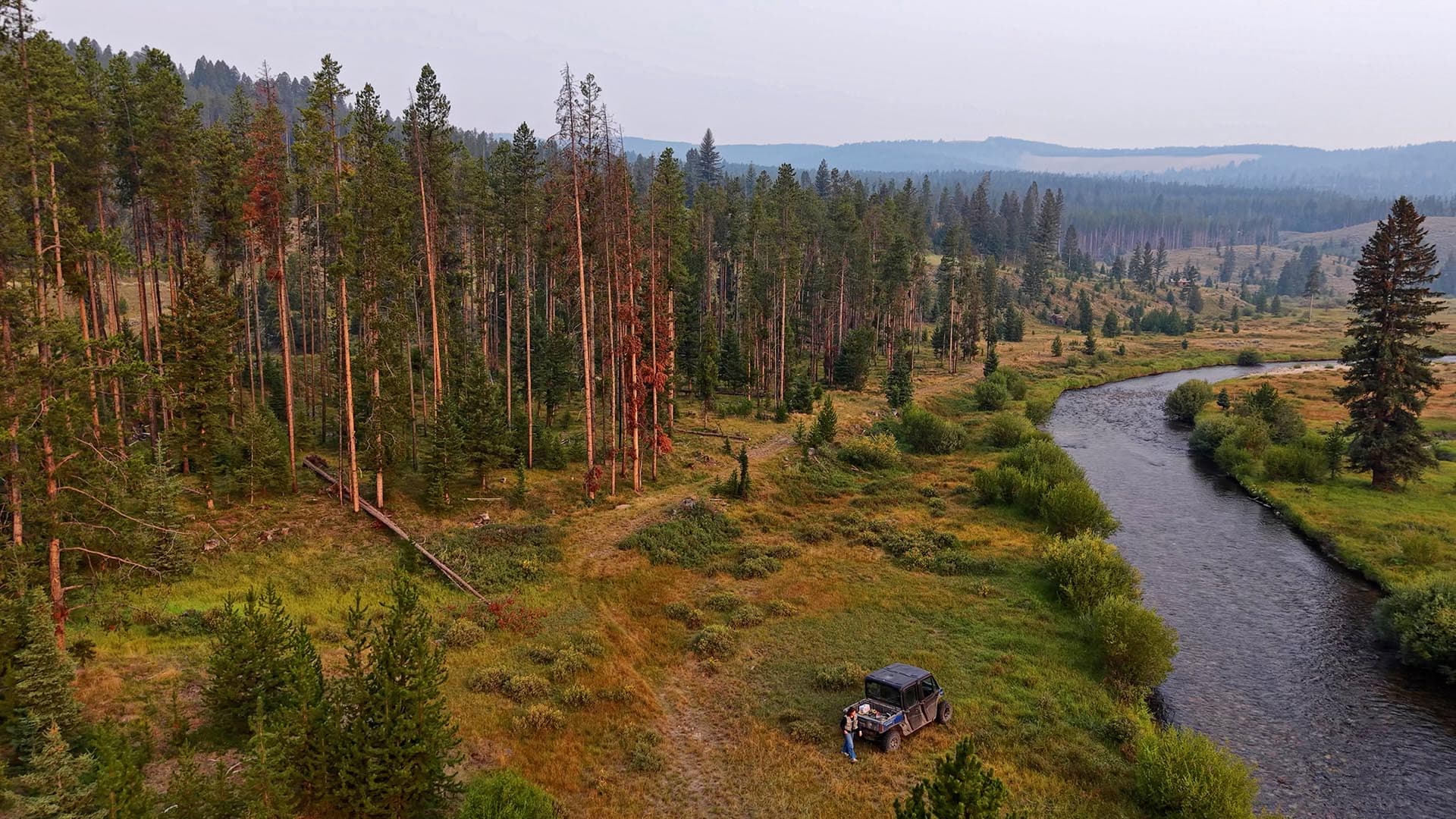Aerial view of a pine forest with a winding river, and two people near a parked off-road vehicle by the shore.