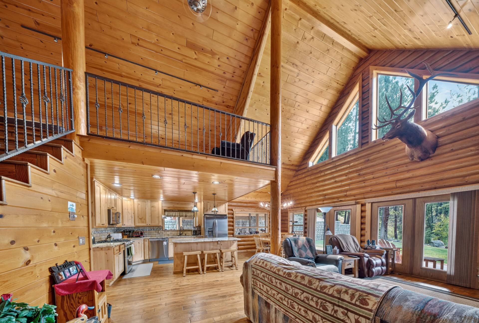 Spacious log cabin interior with a high vaulted wooden ceiling, a kitchen and living area, and a mounted deer head by large windows overlooking trees and a yard.