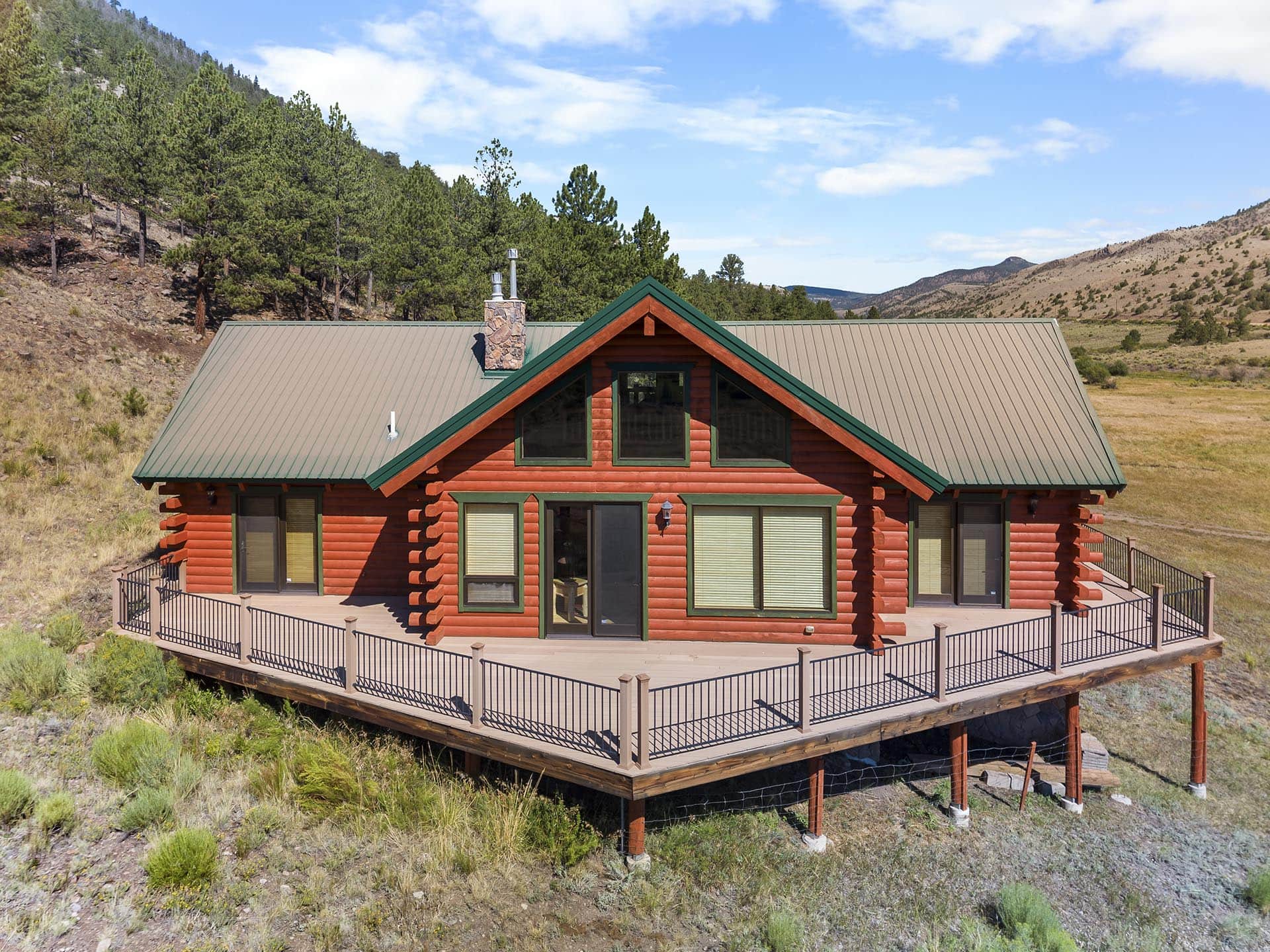 Red log cabin with a green metal roof on a raised deck, set in a grassy, hilly landscape with pine trees and blue sky behind.