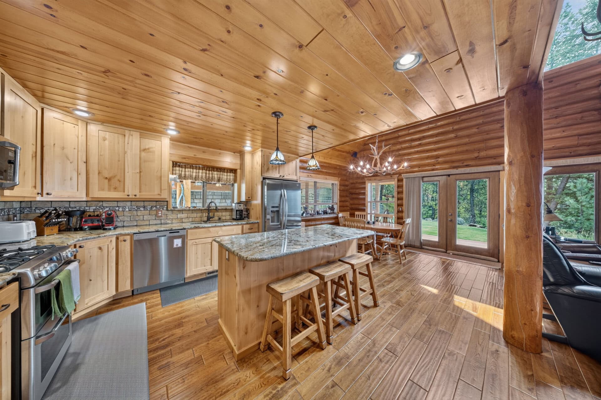 Open-concept rustic kitchen in a log cabin: wood cabinets, island with granite top, stainless appliances, and pendant lights plus a dining area near large windows.