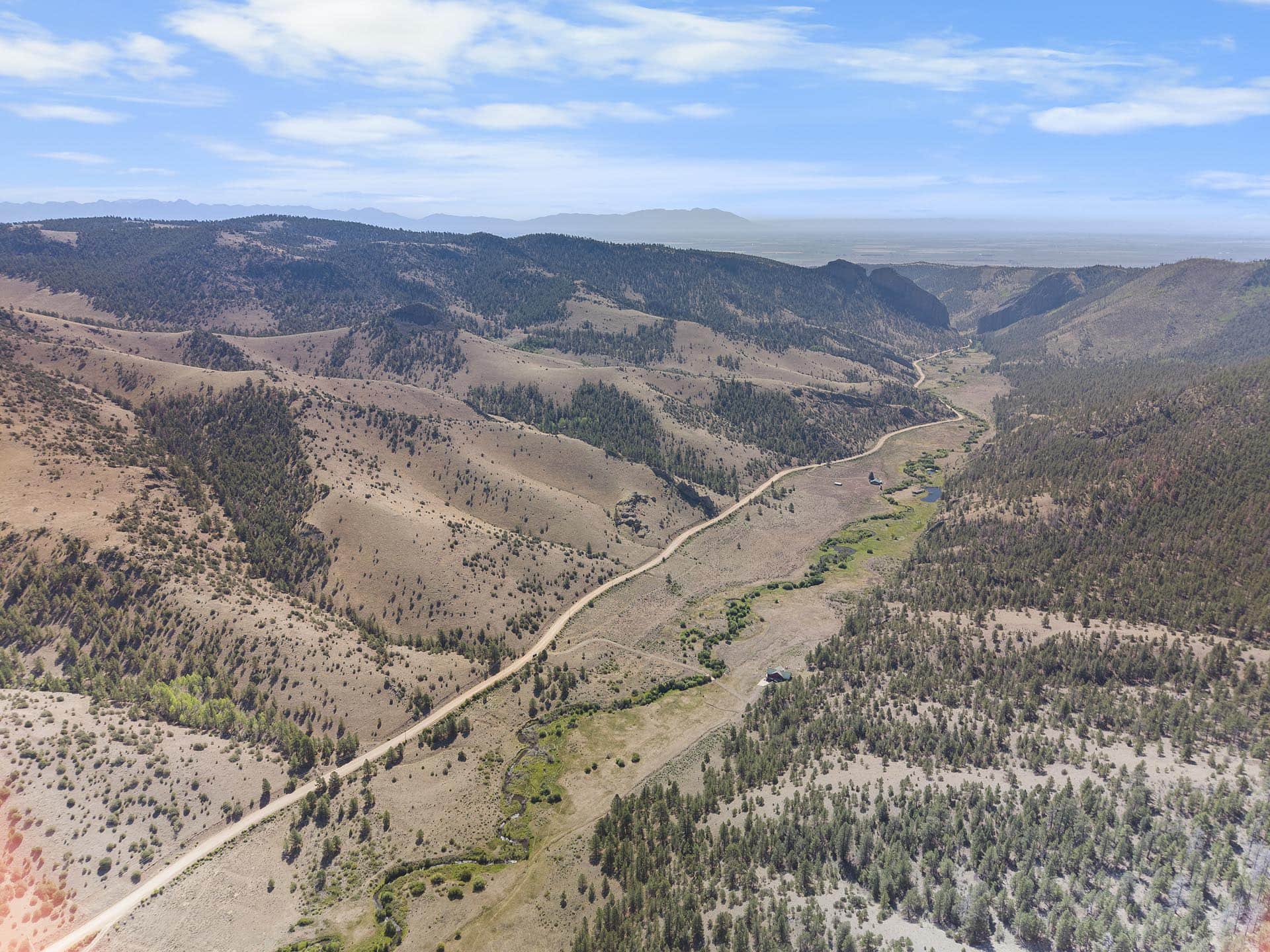 Aerial view of rolling brown hills with scattered trees and a winding dirt road through the valley under a blue sky.