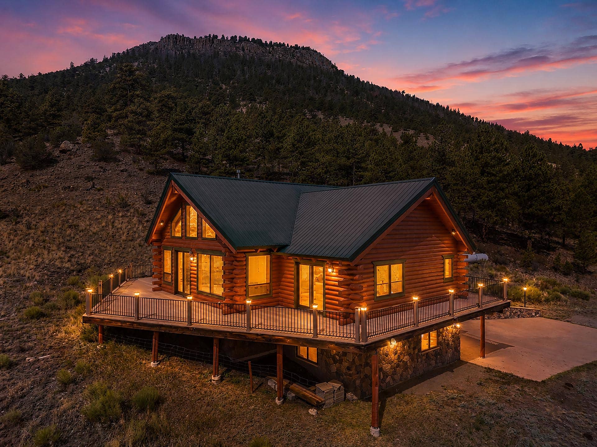 Log cabin with a green roof glowing warmly at dusk, perched on a rocky hillside with pine trees and a sunset sky.