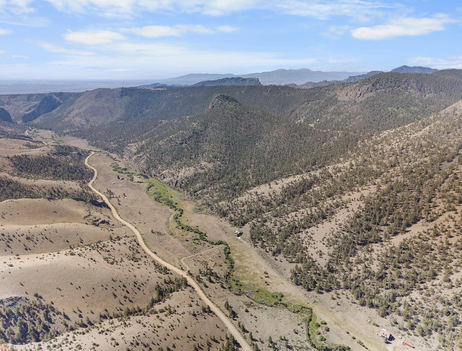 Aerial view of arid mountain landscape with a winding dirt road, scattered trees, and distant blue mountains under a bright sky.