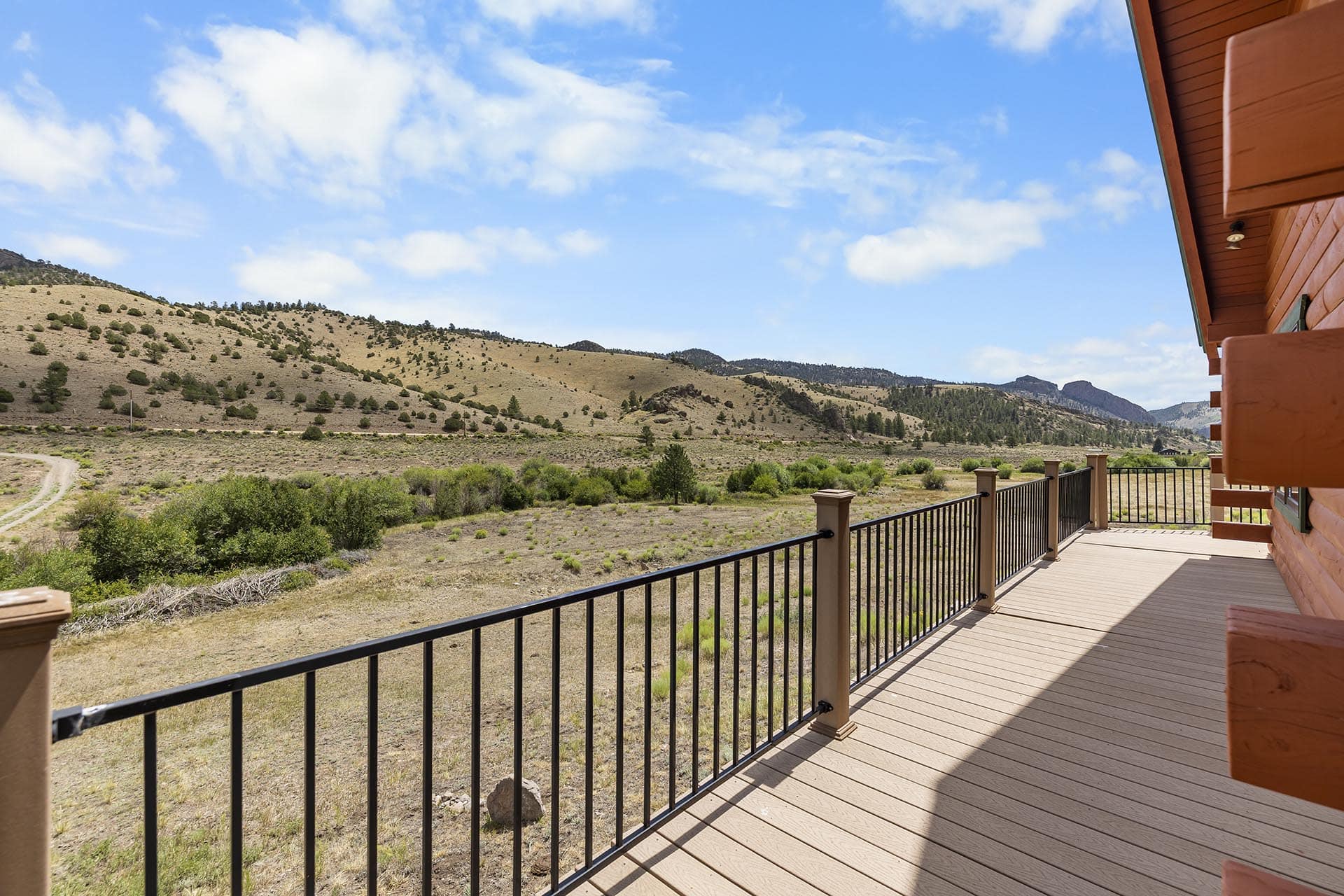 Wood deck with dark metal railing overlooking a dry, grassy hillside and distant mountains on a sunny day. A red wood house edge is on the right.