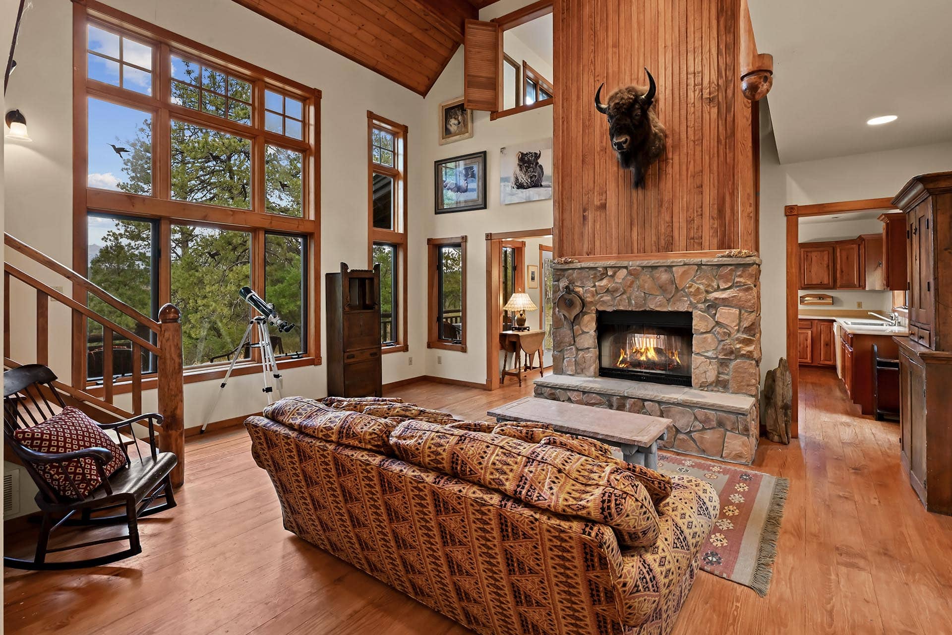 Cozy living room with a stone fireplace, wood-paneled walls, and large windows showing trees outside; buffalo head mounted above the fireplace.