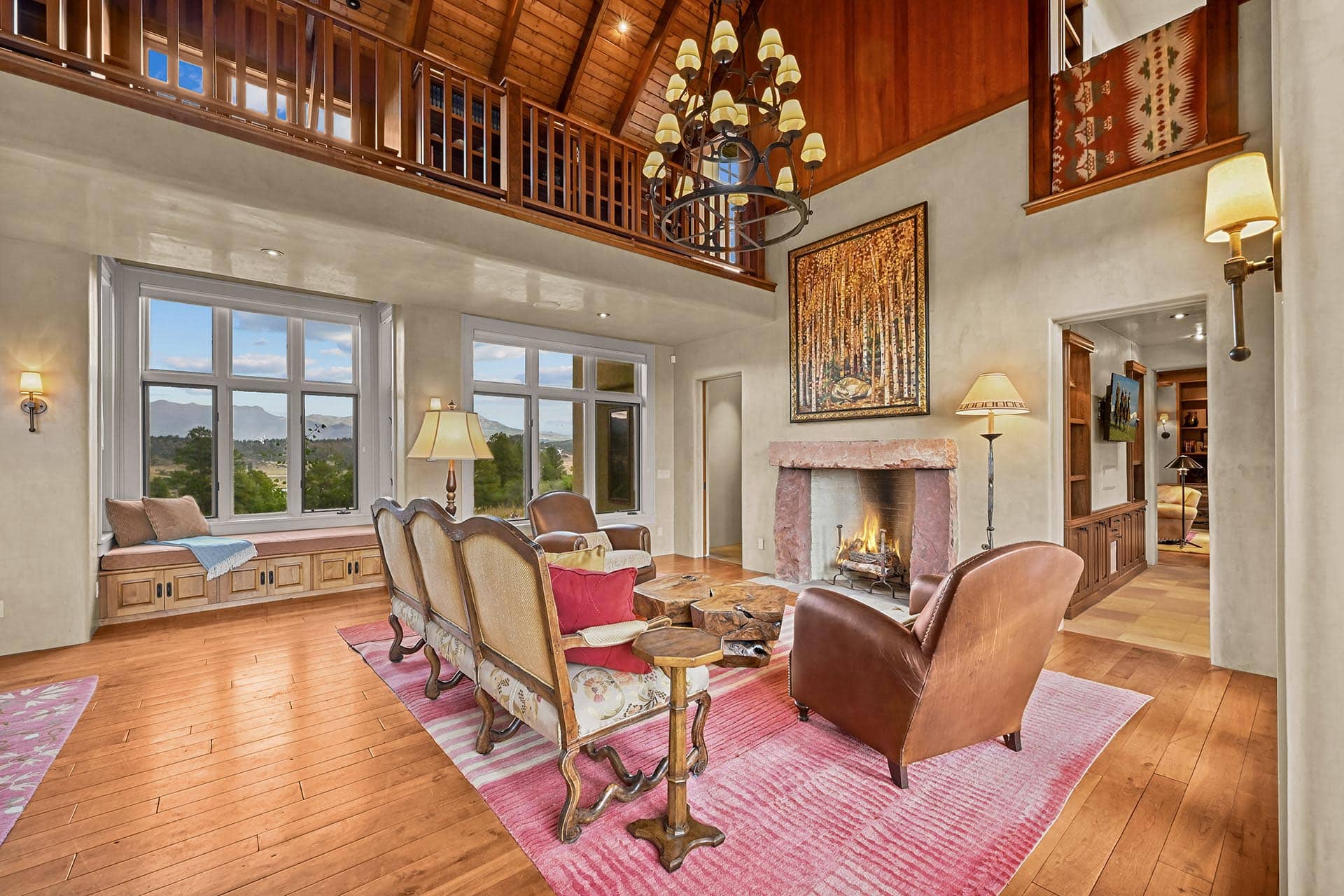 Open-concept living room with a high wooden ceiling, chandelier, stone fireplace, and large windows showing a mountain view.