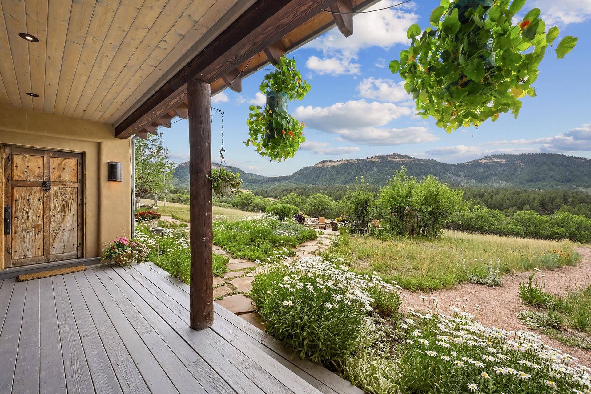 Rustic front porch with weathered wooden door, hanging planters, and a meadowed landscape of hills under a blue sky with clouds.