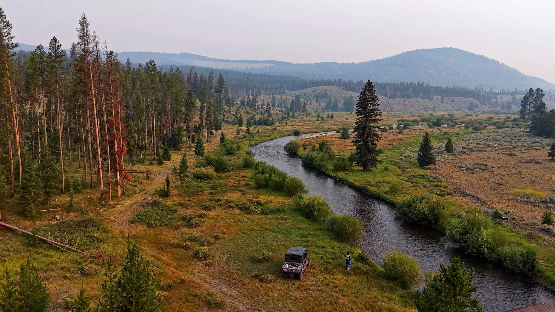 Aerial view of a winding river through a grassy valley with tall pines on the left and distant hills on the horizon, a vehicle and person near the riverbank.