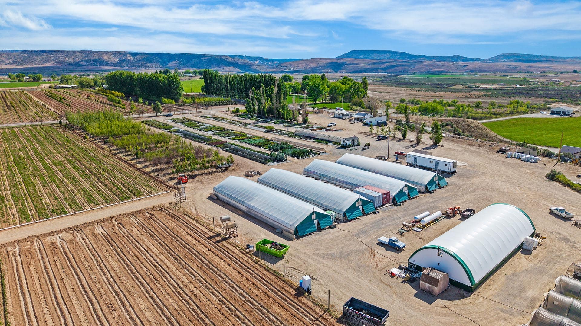 Aerial view of a farm with greenhouses, fields, and distant hills under a blue sky.
