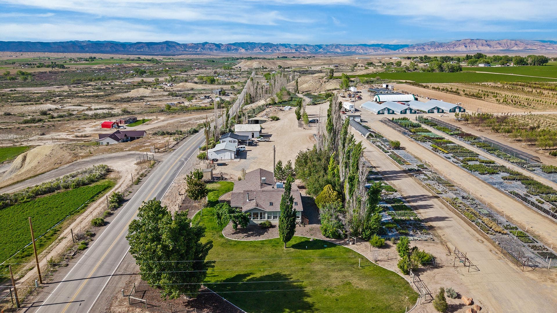Aerial view of a rural valley with houses along a main road, rows of greenhouses, and irrigated fields, mountains on the horizon.
