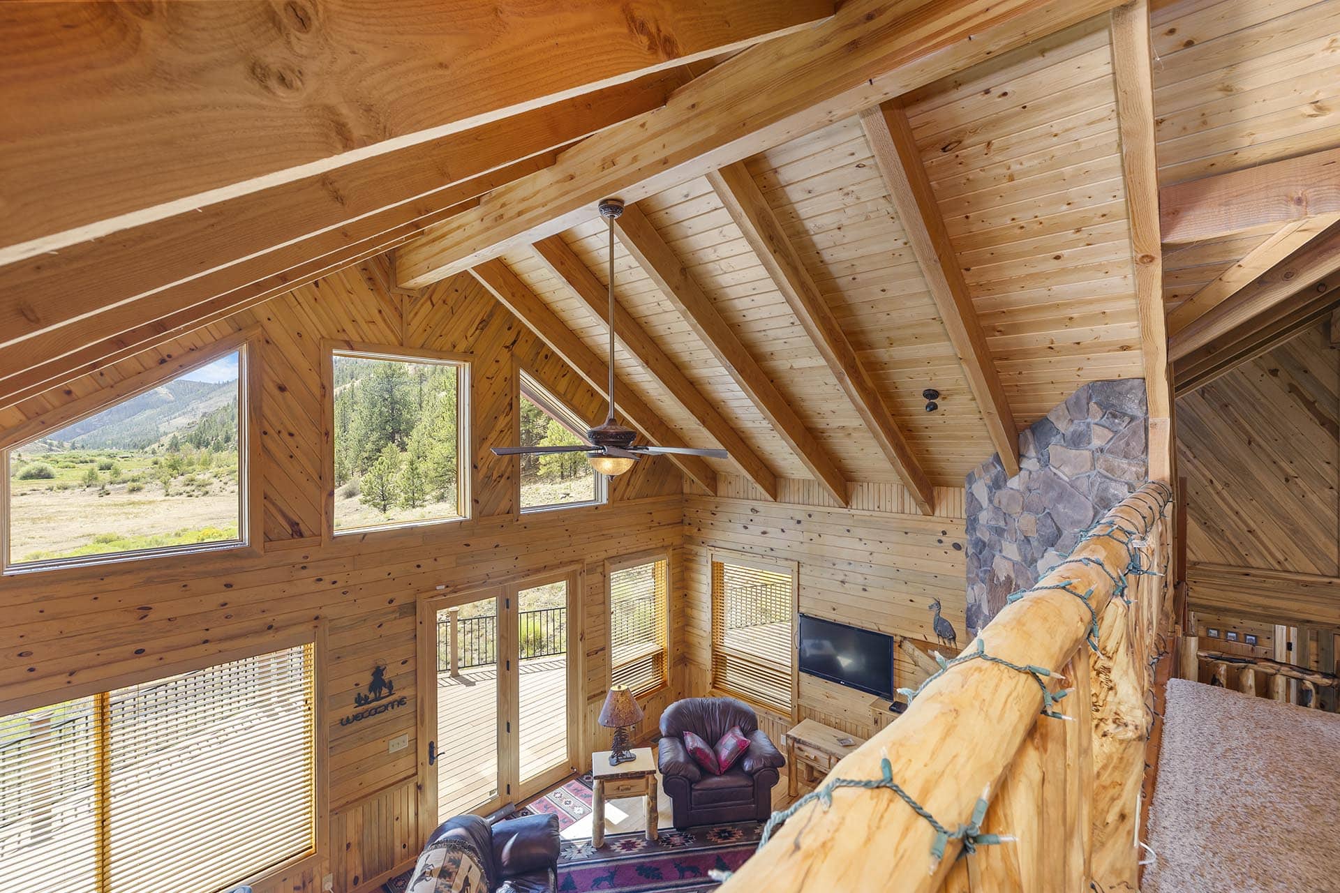 Interior of a rustic log cabin living room with a vaulted wooden ceiling, exposed beams, and large windows showing a mountain landscape outside, plus a stone fireplace to the right.