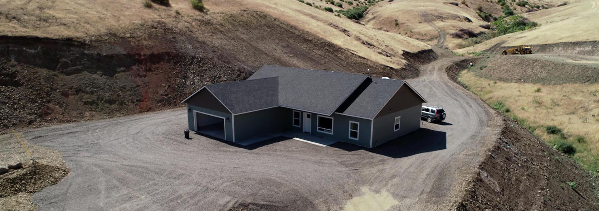 Single-story gray house with an attached garage sits in a dry, excavated landscape with a circular gravel driveway and distant construction equipment.